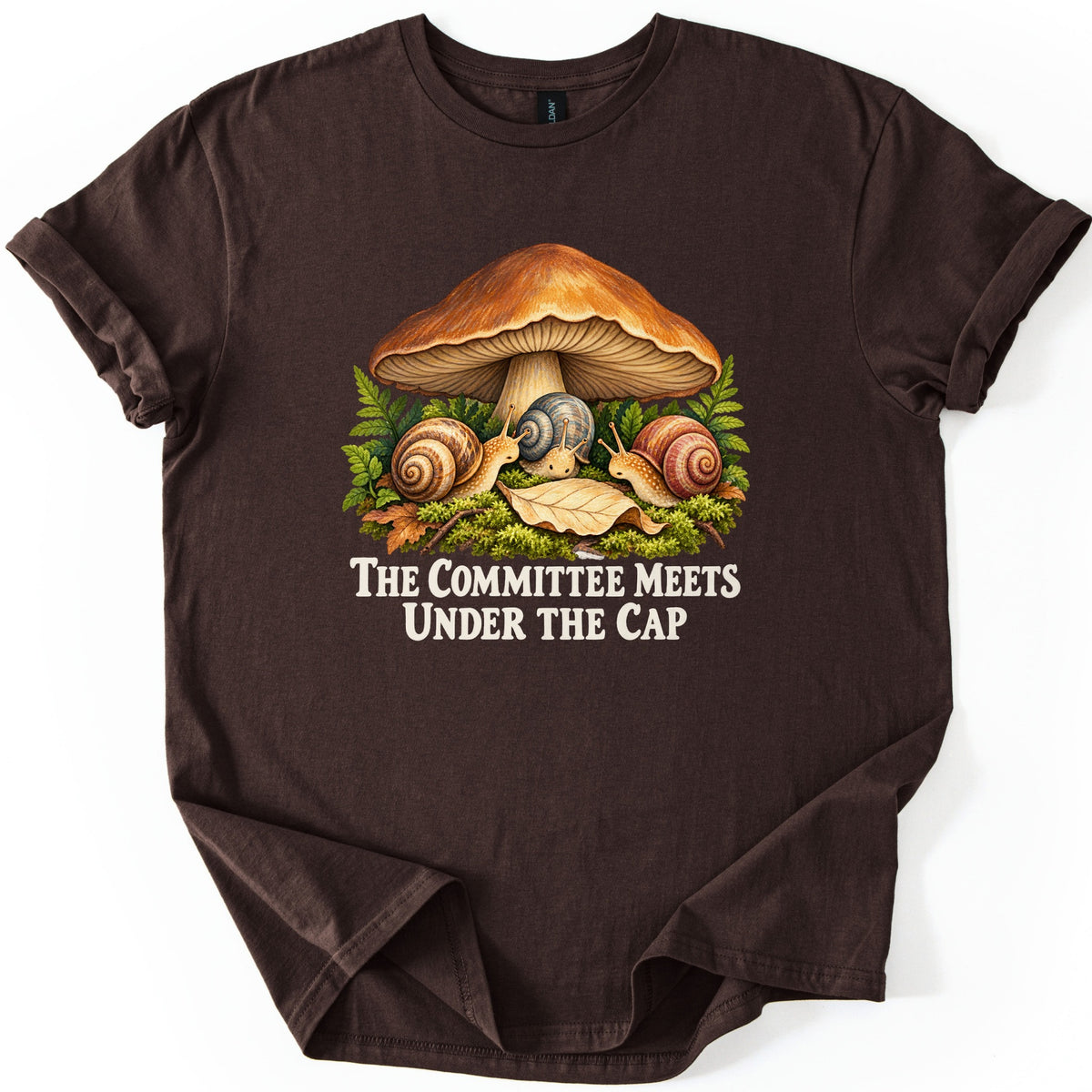 The Committee Meets Under the Cap Comfort Colors T-shirt in Dark Chocolate, laid flat. Three snails gather on moss beneath a large brown mushroom, framed by ferns and a fallen leaf, above cream words The Committee Meets Under the Cap. The shirt lies on a white background with rolled sleeves.