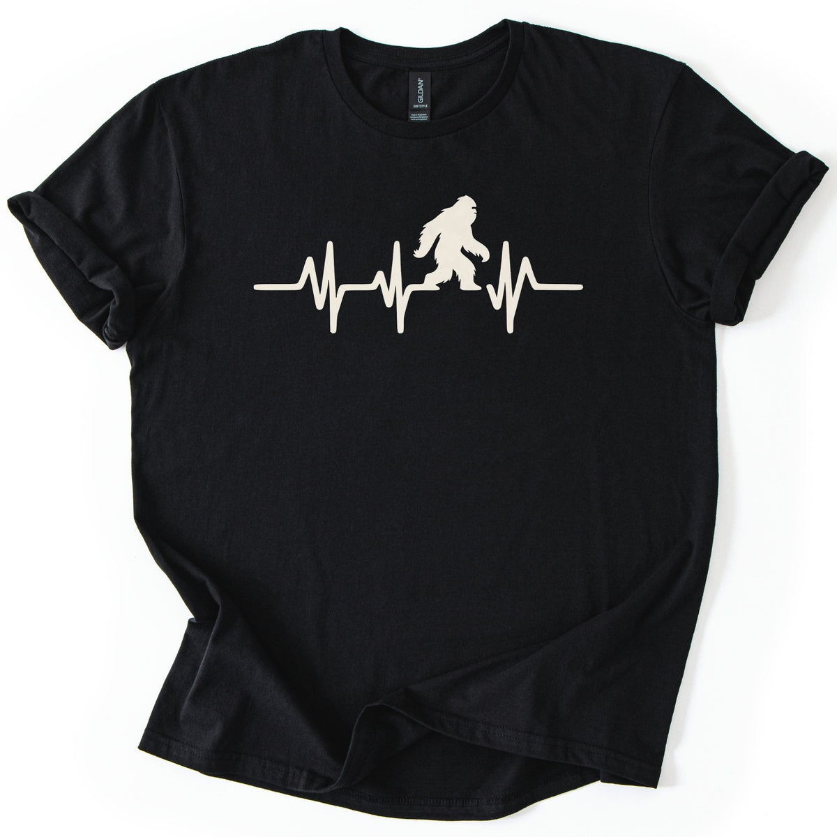 Yeti Heartbeat Comfort Colors T-shirt in Black, laid flat. A cream-colored heartbeat line runs across the chest, with a walking Bigfoot silhouette standing within it; there are no words. The sleeves are cuffed and the shirt lies on a white background.