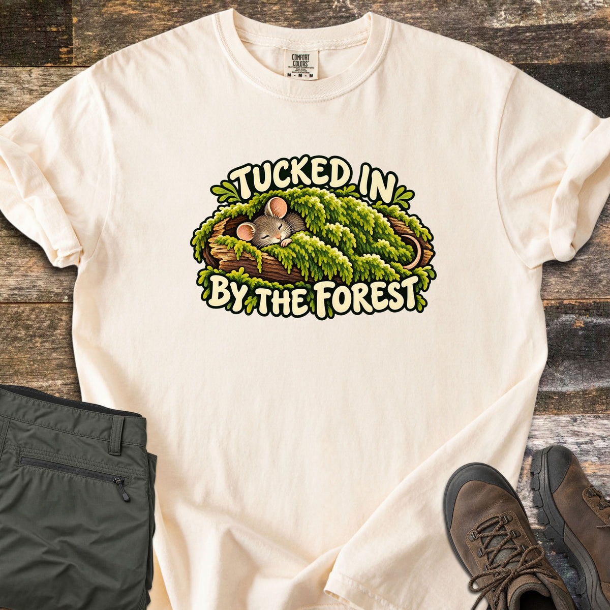 Tucked in by the Forest Comfort Colors T-shirt in Ivory, laid flat. A sleeping cartoon mouse lies under a blanket of green moss in a hollow log between the words Tucked In By The Forest. Folded olive hiking pants and brown hiking boots sit on weathered wood planks.