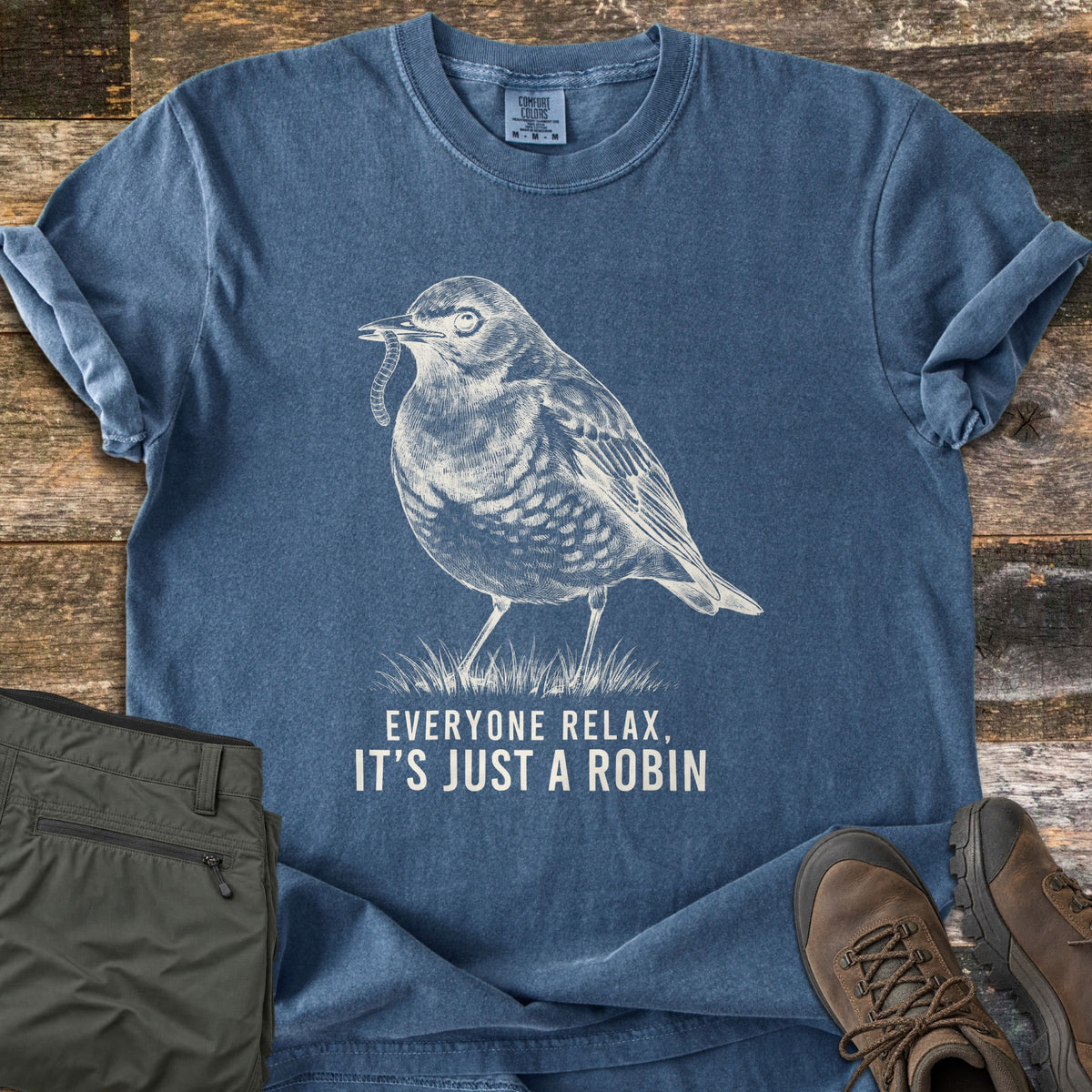 Everyone Relax It's Just A Robin Comfort Colors T-shirt in Denim, laid flat. A white sketch of a robin with a worm in its beak, standing on grass, above the words Everyone Relax, It's Just A Robin. Sleeves rolled, on weathered wood with olive hiking pants and brown hiking boots.