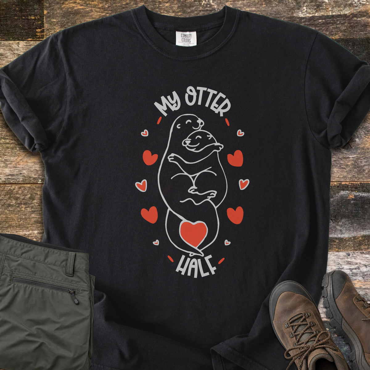 My Otter Half Comfort Colors T-shirt in Black, laid flat. Two smiling cartoon otters hug in white line art, ringed by red hearts with a red heart at their tails, between the words My Otter Half. Folded olive pants and brown hiking boots sit beside it on weathered wood.