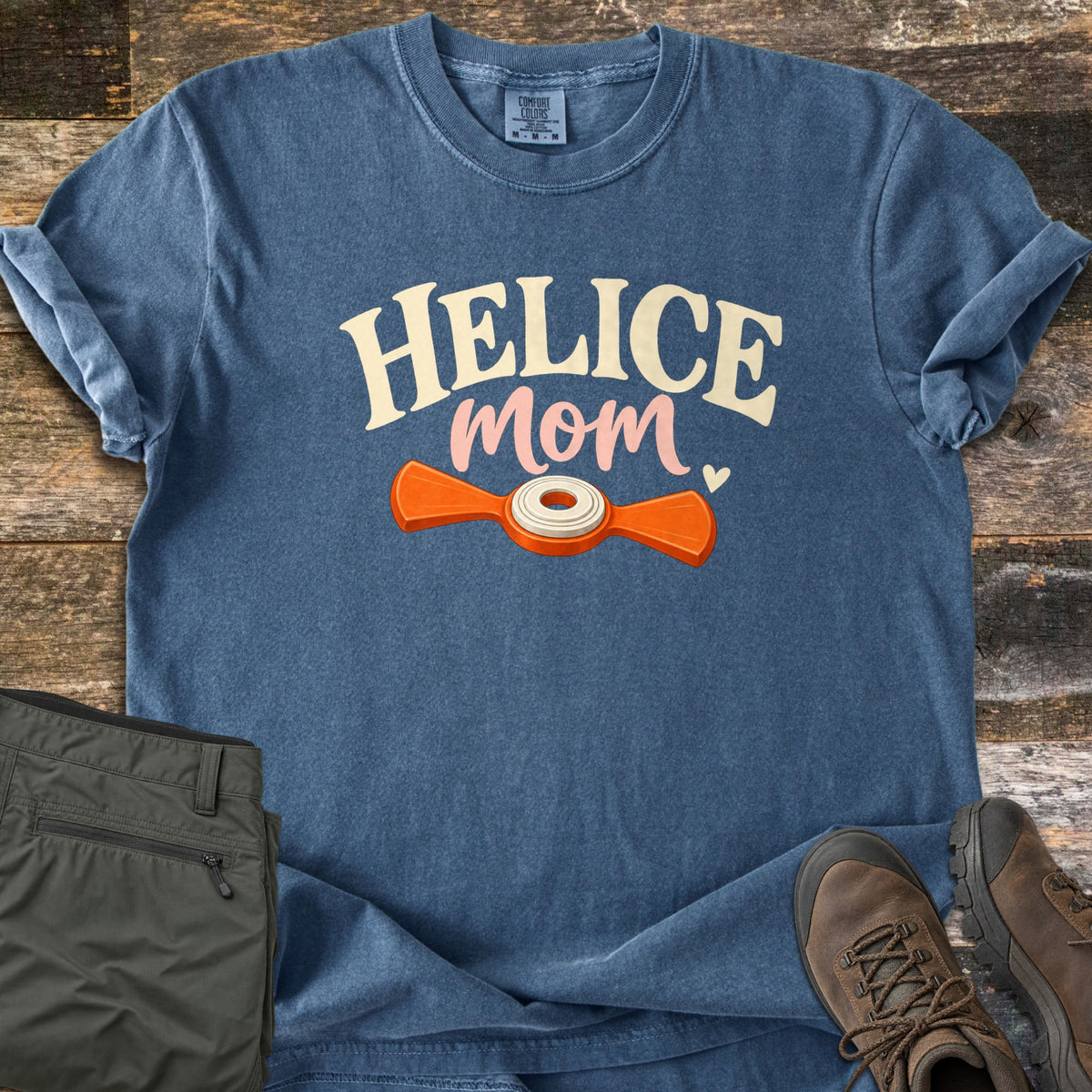 Helice Mom Comfort Colors T-shirt in Denim, laid flat. The word HELICE in cream serif letters over pink script Mom with a small heart, above an orange two-bladed helice target with a white center. Laid on rustic wood with olive cargo pants and brown hiking boots.