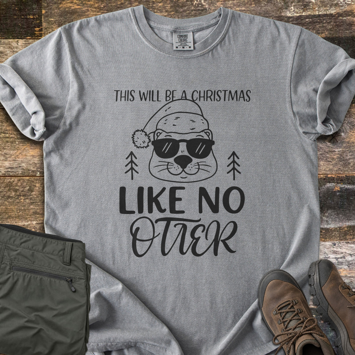 Christmas Like No Otter Comfort Colors T-shirt in Grey, laid flat. A cartoon otter in a Santa hat and sunglasses sits between two pine trees, with This Will Be a Christmas above and Like No Otter below, in black. Olive cargo pants and brown hiking boots lie on weathered wood.