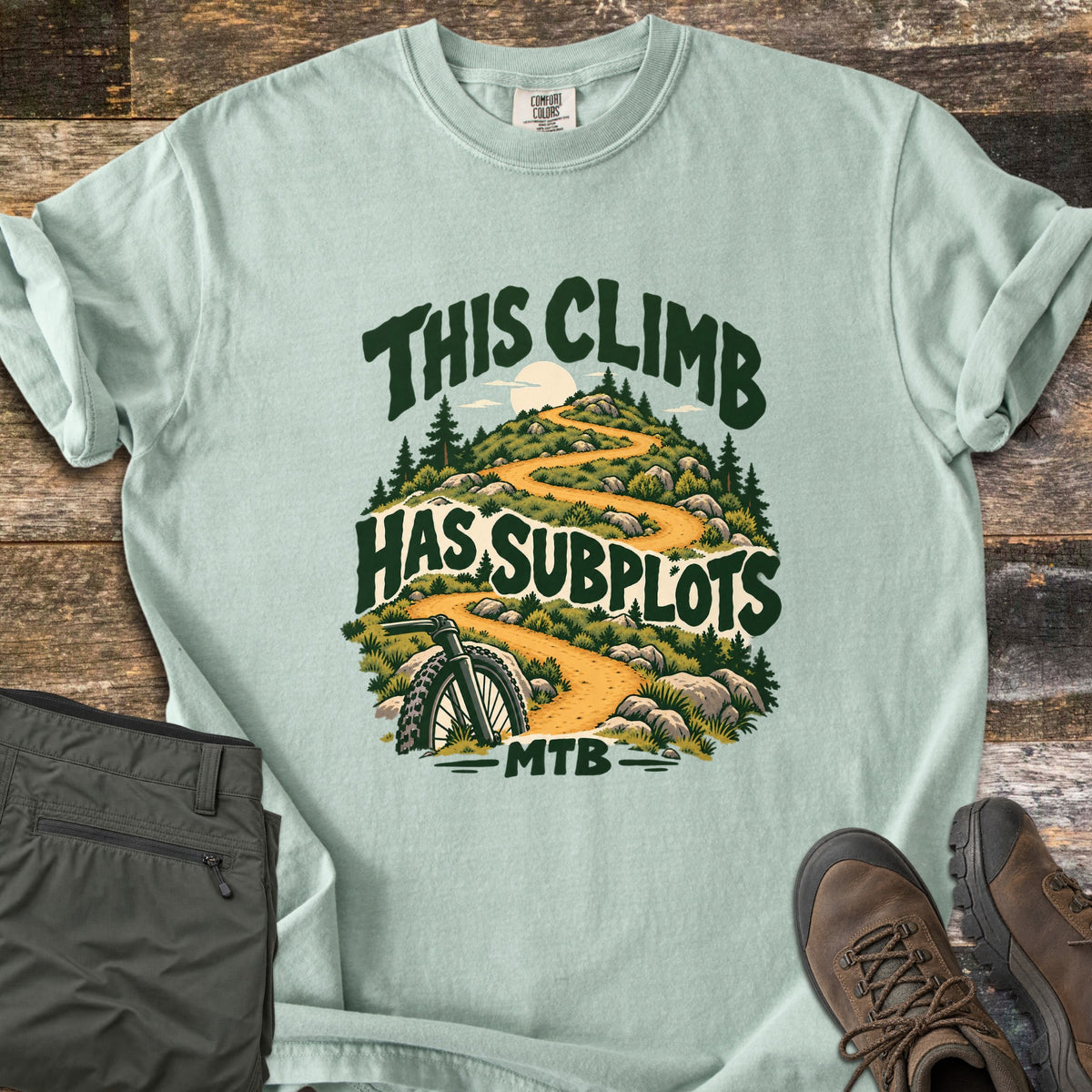 This Climb Has Subplots Comfort Colors T-shirt in Bay, laid flat. Dark green words This Climb Has Subplots MTB wrap around a winding trail up a rocky, pine-dotted hill with a pale sun and a mountain bike wheel. Folded olive pants and brown hiking boots sit on rustic wood.