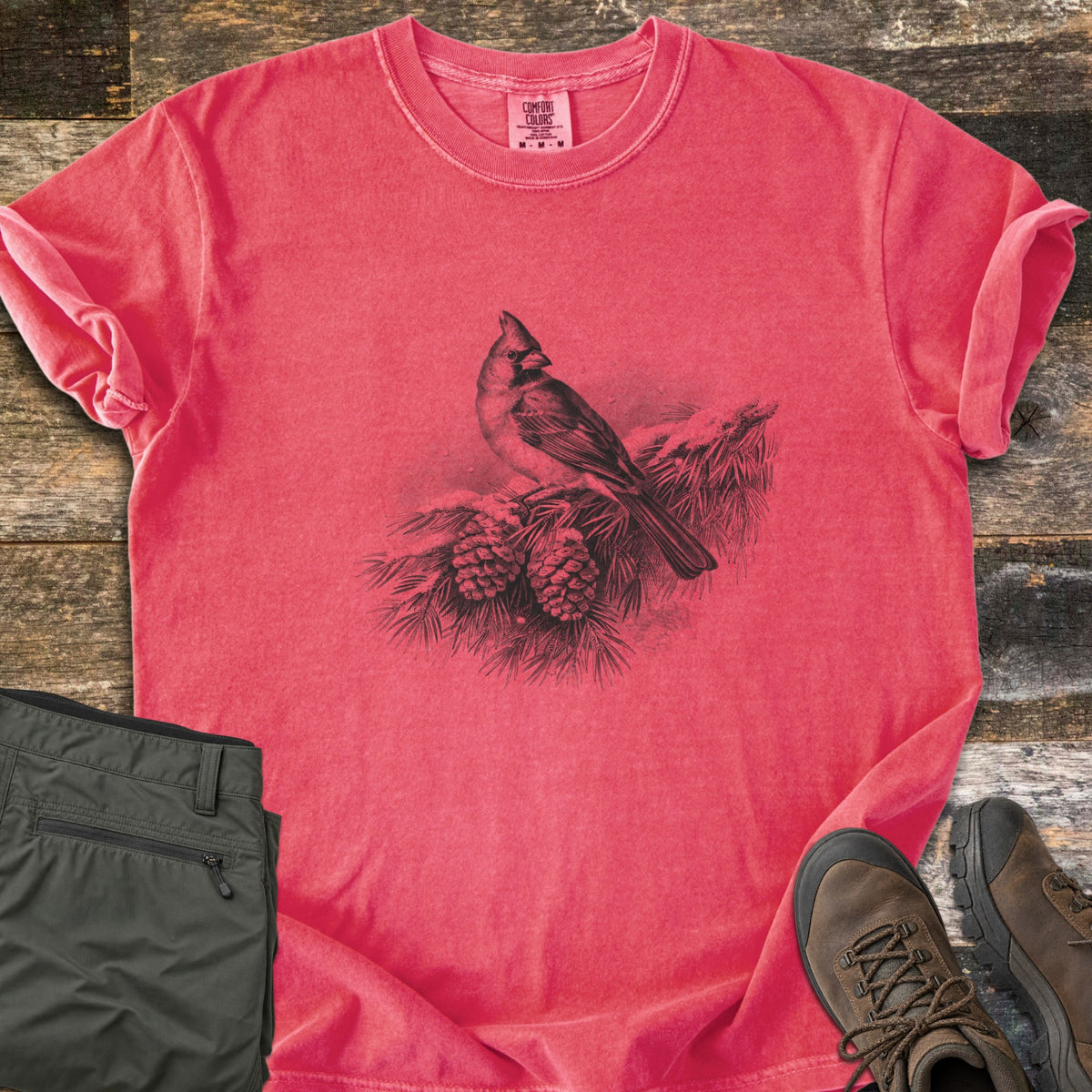 Cardinal On A Snowy Bough Comfort Colors T-shirt in Watermelon, laid flat. A grayscale drawing of a cardinal perched on a snow-covered pine branch with two pinecones, with no words. Sleeves rolled, on weathered wood planks beside olive hiking pants and brown hiking boots.