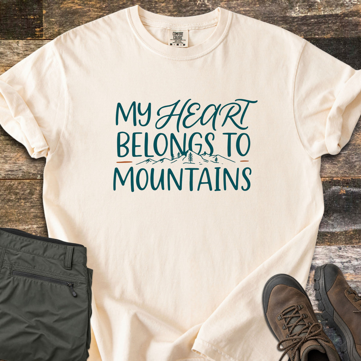 My Heart Belongs to Mountains Comfort Colors T-shirt in Ivory, laid flat. Dark teal lettering reads My Heart Belongs to Mountains, with Heart in script above sketched peaks and pine trees flanked by rust-orange dashes. Olive hiking pants and brown boots rest on rustic wood.