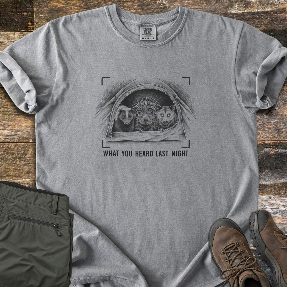 What You Heard Last Night Comfort Colors T-shirt in Grey, laid flat. A black sketch of a skunk, porcupine and opossum peeking out of an unzipped tent, framed by camera brackets, above the words What You Heard Last Night. Olive hiking pants and brown boots lie on weathered wood.