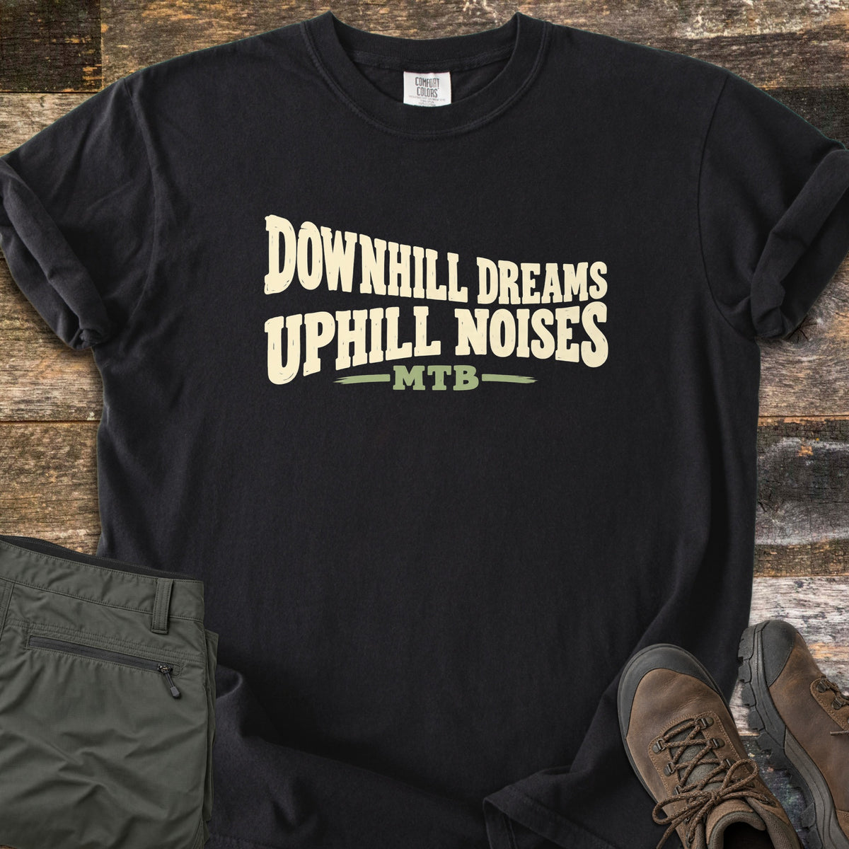 Downhill Dreams Uphill Noises Comfort Colors T-shirt in Black, laid flat. Cream wavy block letters read Downhill Dreams Uphill Noises above MTB in green between two green lines. Sleeves rolled on weathered wood planks, with folded olive hiking pants and brown leather hiking boots.