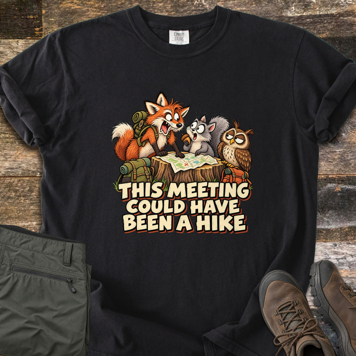 This Meeting Could Have Been a Hike Comfort Colors T-shirt in Black, laid flat. A shouting cartoon fox, a squirrel holding an acorn and a sleepy owl study a trail map on a tree stump above the words This Meeting Could Have Been a Hike. Olive pants and brown hiking boots lie on dark wood.