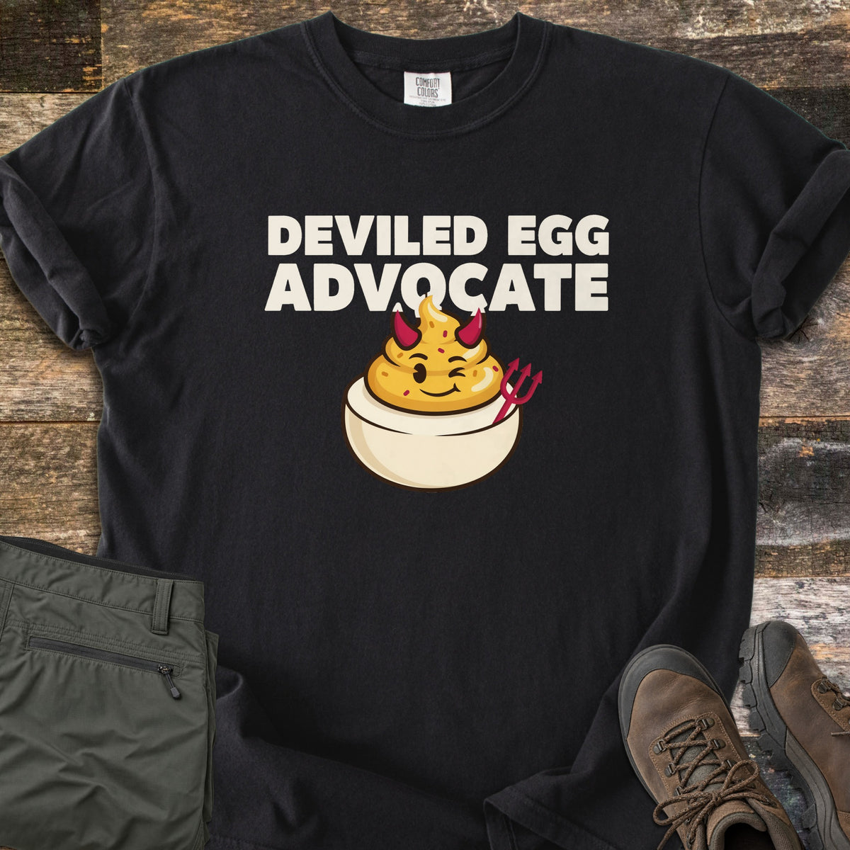 Deviled Egg Advocate Comfort Colors T-shirt in Black, laid flat. Cream letters read Deviled Egg Advocate above a winking cartoon deviled egg with red devil horns and a red pitchfork. Folded olive hiking pants and brown hiking boots sit on weathered wood planks.
