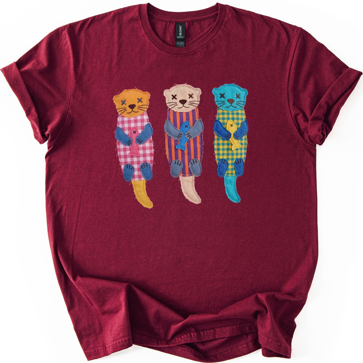 Patchwork Otter Comfort Colors T-shirt in Maroon, laid flat. Three fabric-patch otters with blue paws and stitched X eyes hug fish: orange in pink gingham, beige in purple and orange stripes, turquoise in yellow gingham. No words. Cuffed sleeves on a white surface.