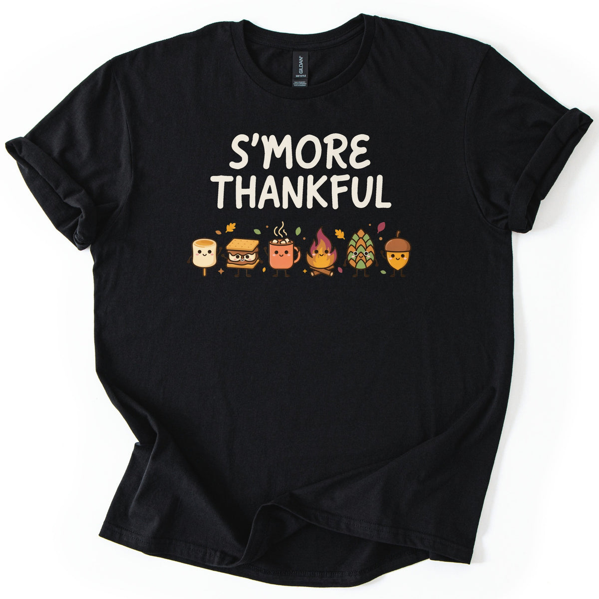 Smore Thankful Comfort Colors T-shirt in Black, laid flat. Cream letters read S'MORE THANKFUL above a row of smiling cartoon characters: a marshmallow, a s'more, a cocoa mug, a campfire, a pinecone and an acorn among autumn leaves. Sleeves rolled, on white.