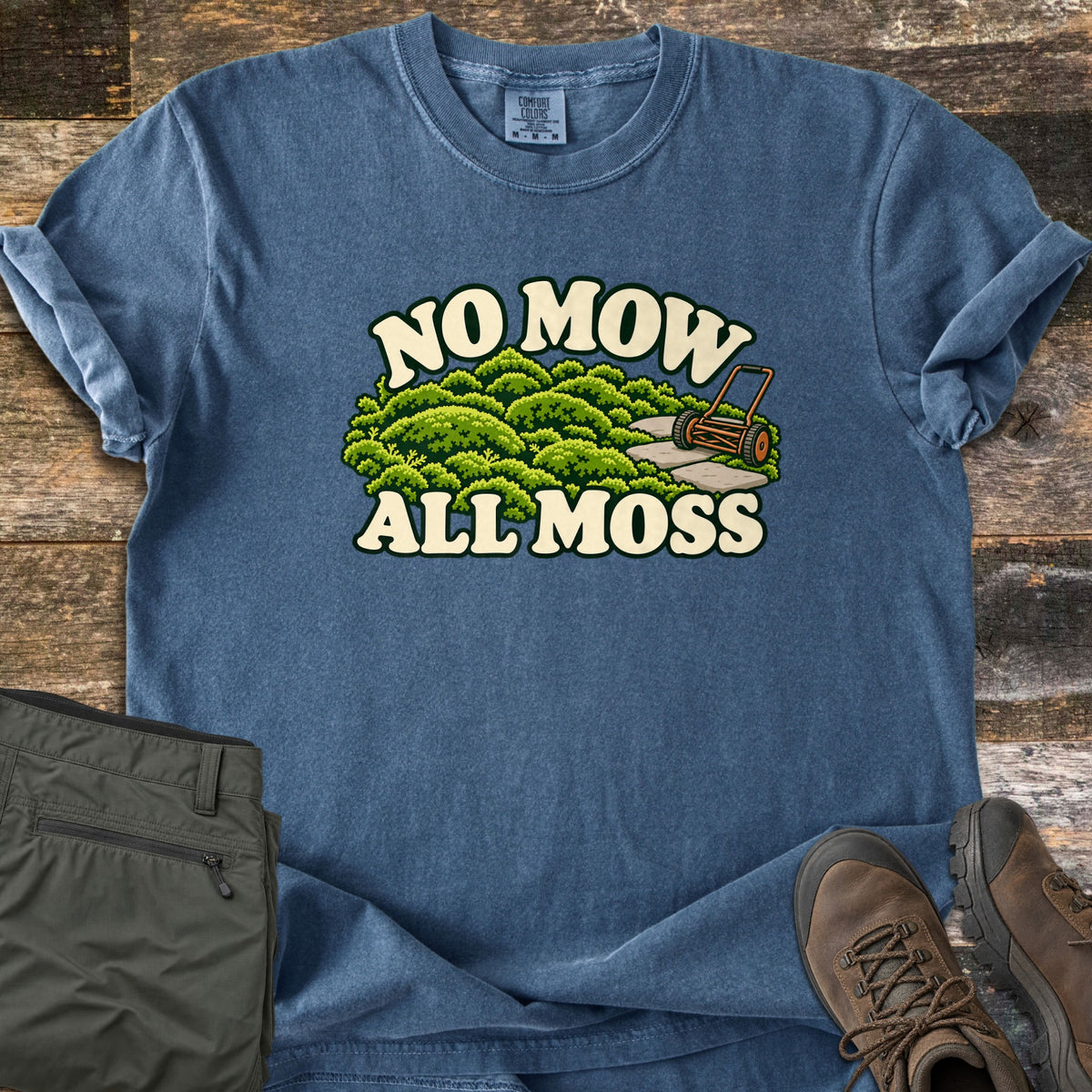 No Mow All Moss Comfort Colors T-shirt in Denim, laid flat. Illustrated mounds of green moss with stepping stones and an old push reel mower, between the words No Mow All Moss. Sleeves rolled, on weathered wood beside olive pants and brown hiking boots.