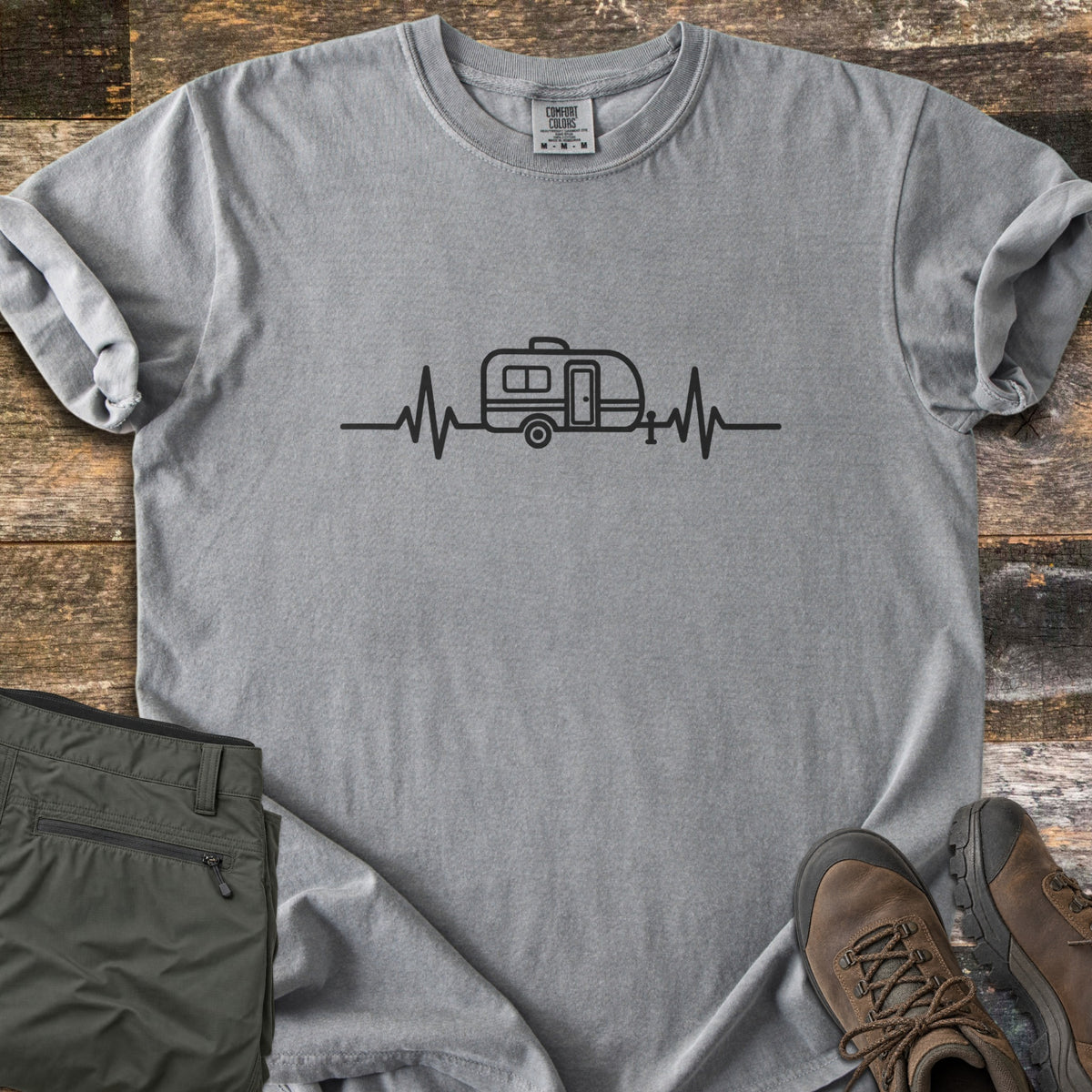 Camper Heartbeat Comfort Colors T-shirt in Grey, laid flat. A black line drawing of a teardrop camper trailer sits in the middle of a heartbeat line across the chest. Folded olive pants and brown hiking boots rest on a rustic wood background.