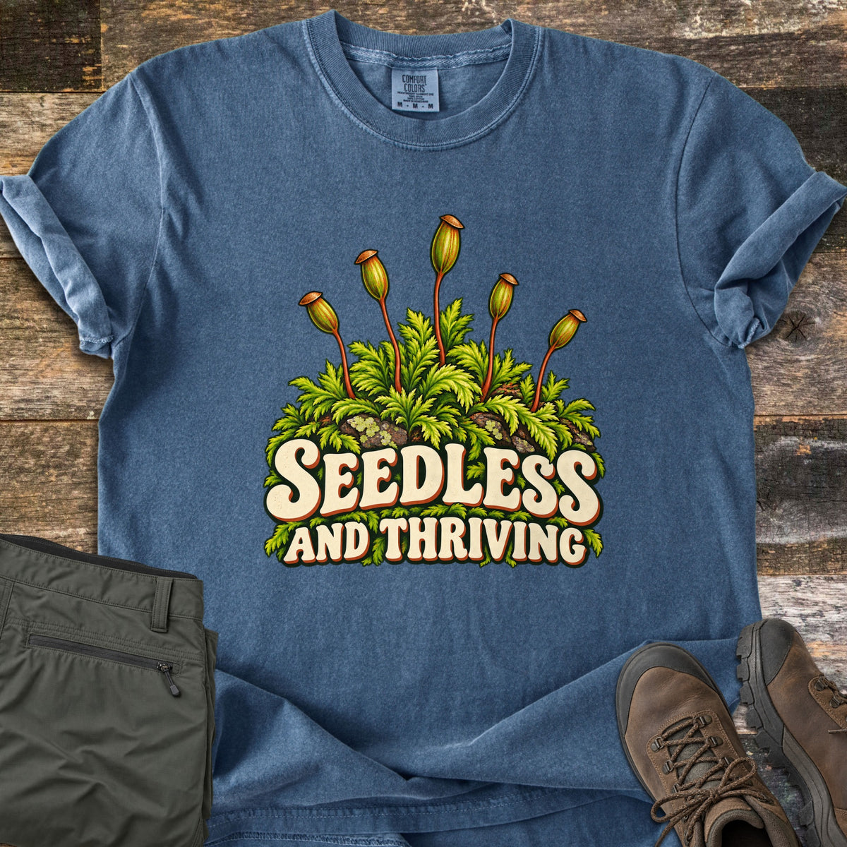 Seedless and Thriving Comfort Colors T-shirt in Denim, laid flat. Moss with tall orange-capped spore stalks and green fronds sits above the words Seedless and Thriving in cream letters. Olive pants and brown hiking boots lie beside it on weathered wood planks.