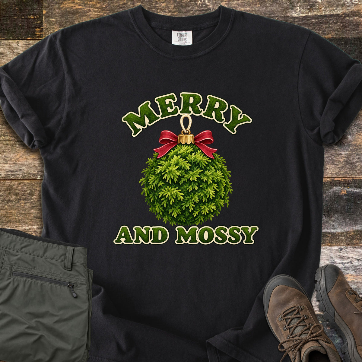 Merry and Mossy Comfort Colors T-shirt in Black, laid flat. A round Christmas ornament made of green moss, topped with a gold cap and red bow, sits between the words Merry and Mossy in green lettering. Olive hiking pants and brown hiking boots lie beside it on weathered wood.