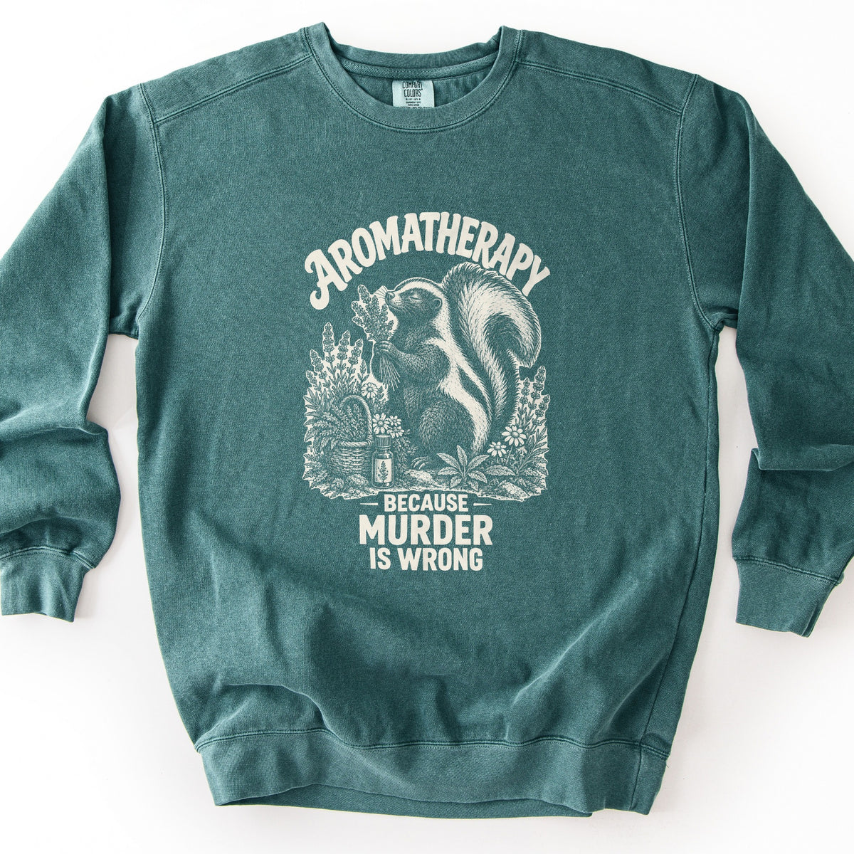 Aromatherapy Because Murder Is Wrong Comfort Colors crewneck sweatshirt in Blue Spruce, laid flat. Cream line art of a skunk sniffing a bundle of lavender among wildflowers, a basket and an essential oil bottle, with Aromatherapy above and Because Murder Is Wrong below. Shown on a white surface.