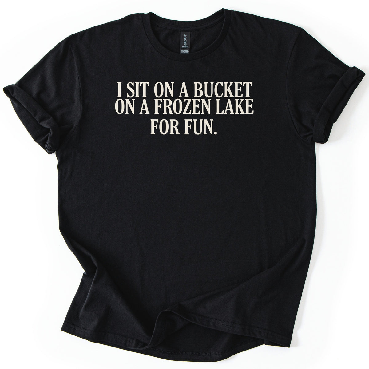 Bucket On A Frozen Lake Comfort Colors T-shirt in Black, laid flat. Cream serif capital letters across the chest read I SIT ON A BUCKET ON A FROZEN LAKE FOR FUN. The shirt lies on a white surface with its sleeves rolled and the hem loosely folded.