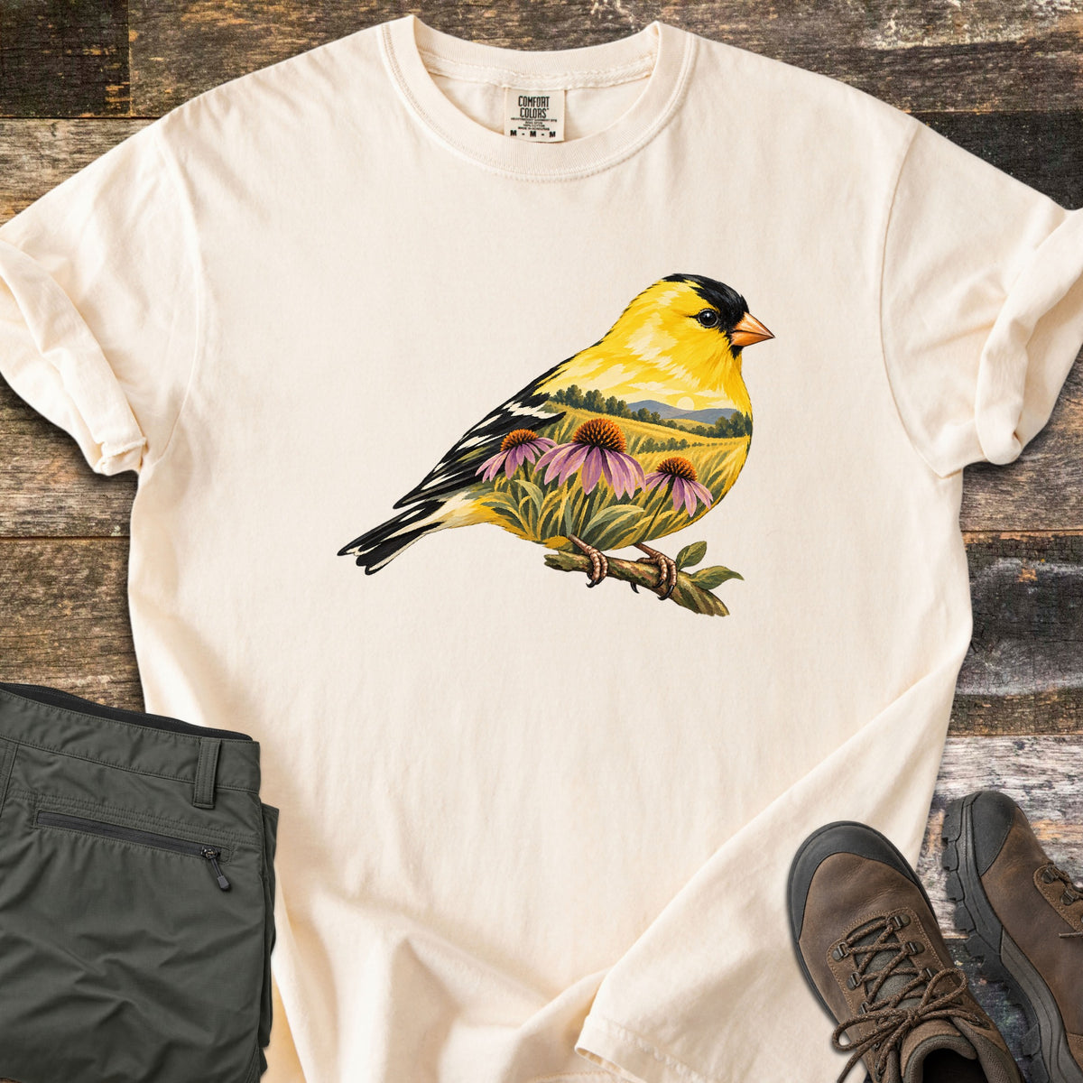 Goldfinch Meadow Comfort Colors T-shirt in Ivory, laid flat. A goldfinch perched on a leafy twig, its body filled with purple coneflowers, tall grass, green hills and a setting sun. No text. Folded olive cargo pants and brown hiking boots rest on rustic planks.