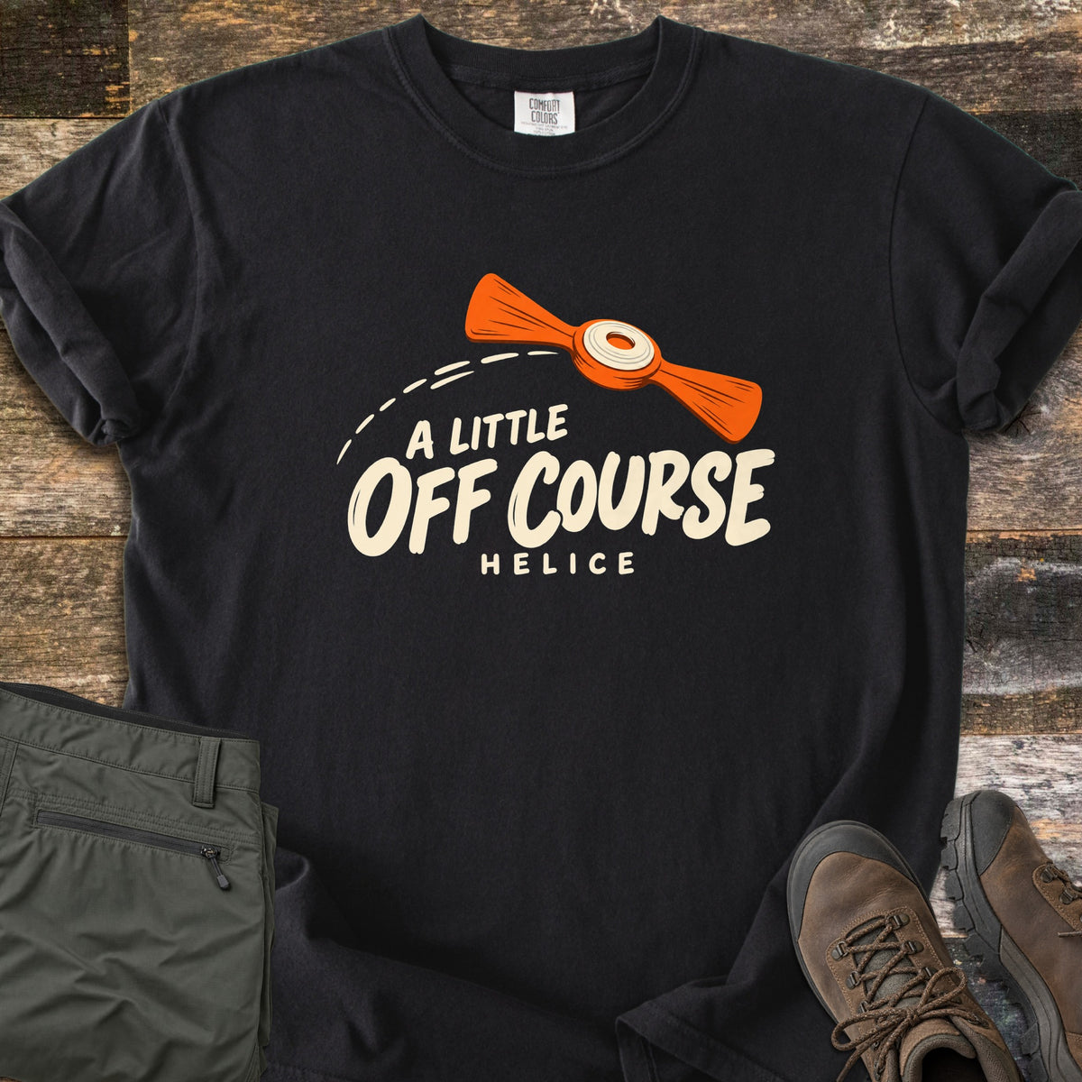 A Little Off Course Comfort Colors T-shirt in Black, laid flat. An orange helice target with a white center flies along a dashed arc above cream text reading A Little Off Course and Helice. Folded olive hiking pants and brown hiking boots sit beside it on weathered wood.