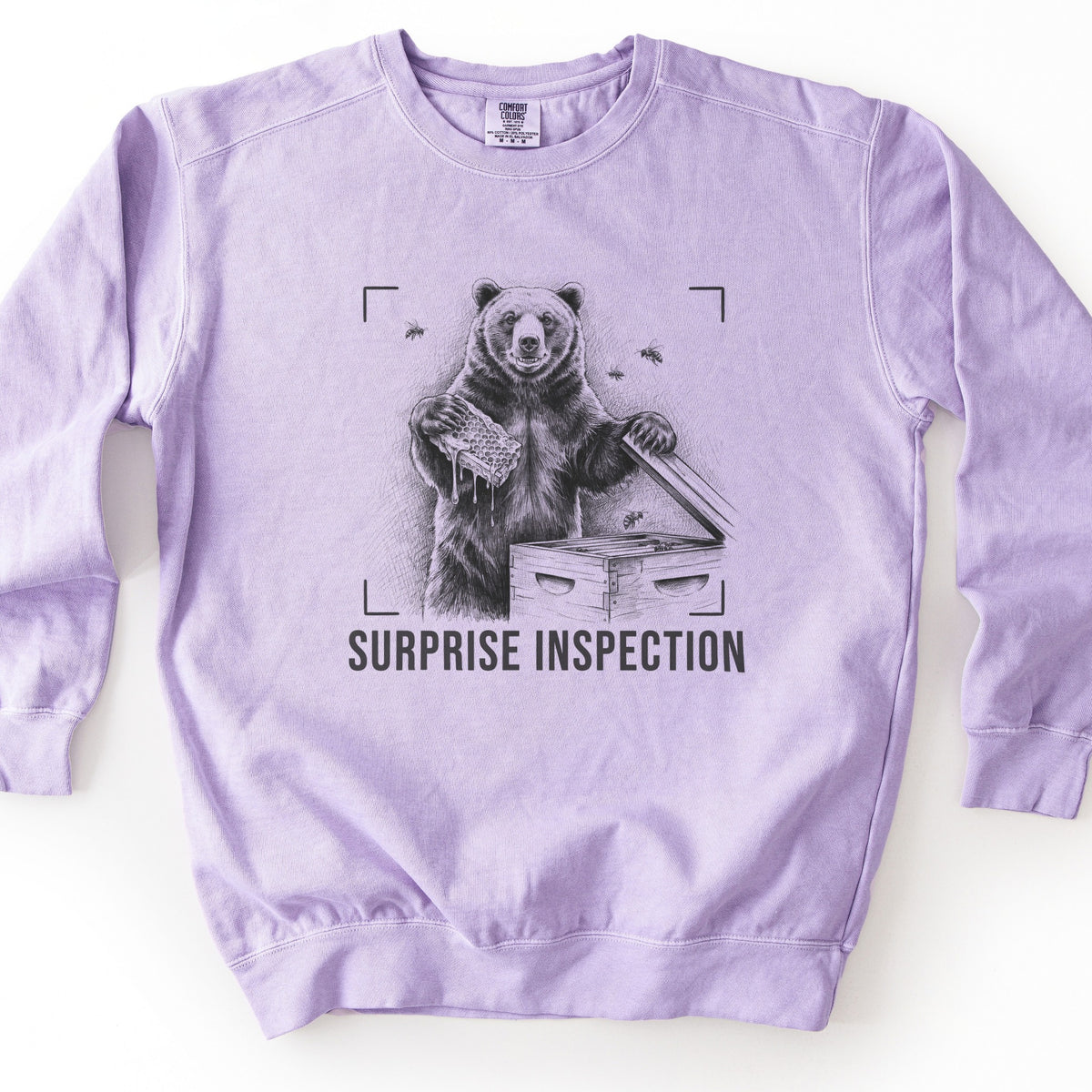 Surprise Inspection Comfort Colors crewneck sweatshirt in Orchid, laid flat. A sketched bear holds a dripping honeycomb and lifts the lid of a wooden beehive as bees fly nearby, inside camera viewfinder corners above the words SURPRISE INSPECTION in black. Laid on a white surface.