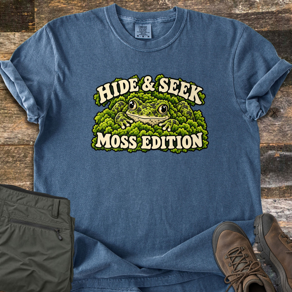 Hide & Seek Moss Edition Comfort Colors T-shirt in Denim, laid flat. A moss-covered frog peeks out from green moss between the words Hide & Seek and Moss Edition. Olive hiking pants and brown hiking boots lie beside it on weathered wood planks.