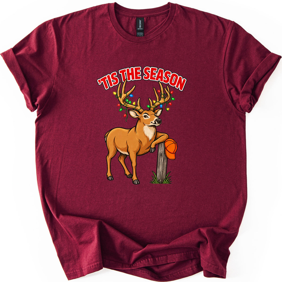 Buck Tis The Season Comfort Colors T-shirt in Maroon, laid flat. Red arched text reads 'TIS THE SEASON above a cartoon buck with colored Christmas lights strung on its antlers, leaning on a wooden post that holds an orange hunting cap. Sleeves rolled, on a white surface.