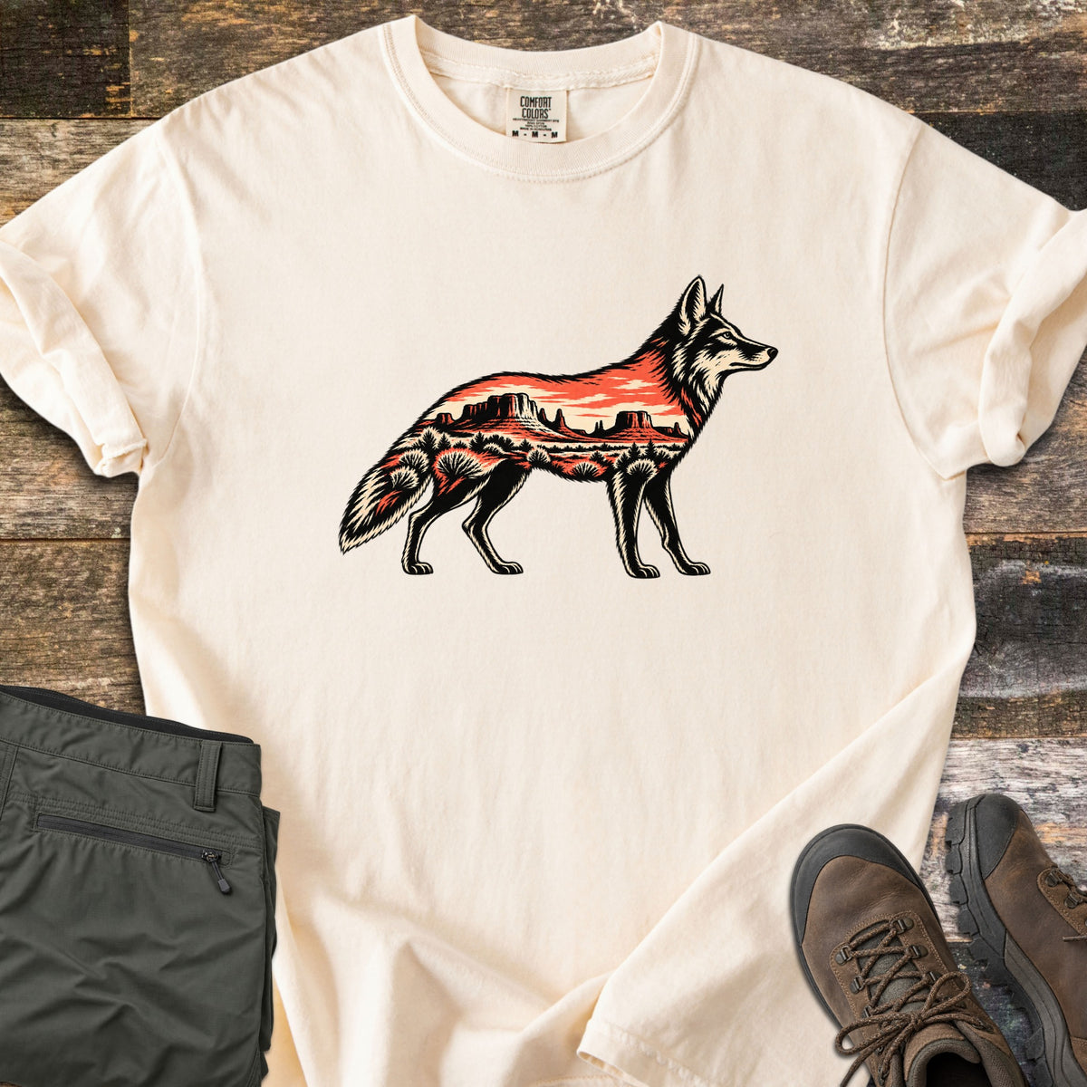 Desert Mesa Coyote Comfort Colors T-shirt in Ivory, laid flat. A side-view coyote silhouette filled with a red desert mesa landscape, sunset sky and shrubs, with no words. Sleeves rolled on rustic wood, beside folded olive hiking pants and brown hiking boots.