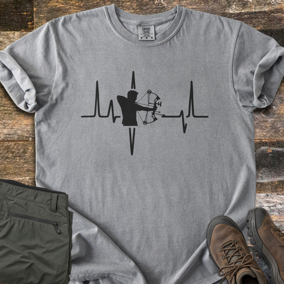 Compound Bow Heartbeat Comfort Colors T-shirt in Grey, laid flat. A black heartbeat line with the silhouette of an archer drawing a compound bow at its center. Sleeves are rolled on a wood plank floor, with olive cargo pants and brown hiking boots beside it.
