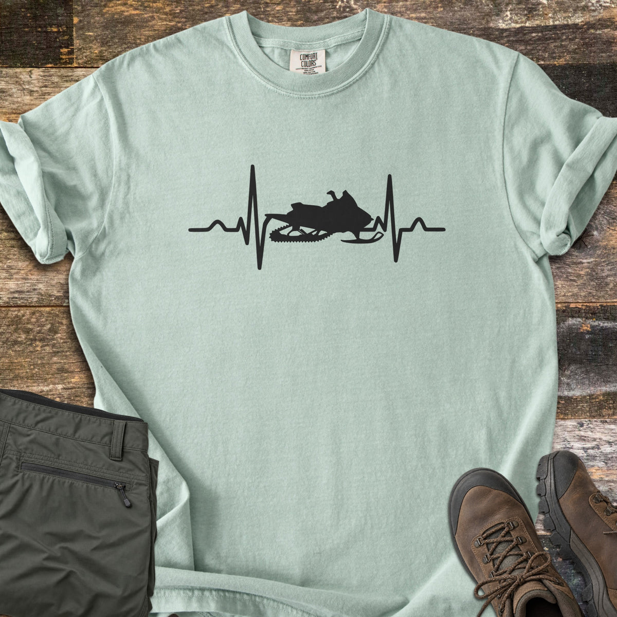 Snowmobile Heartbeat Comfort Colors T-shirt in Bay, laid flat. A black heartbeat line runs across the chest of the pale green shirt with a snowmobile silhouette in its center, with no words. Sleeves are rolled, on wood planks beside folded olive pants and brown hiking boots.