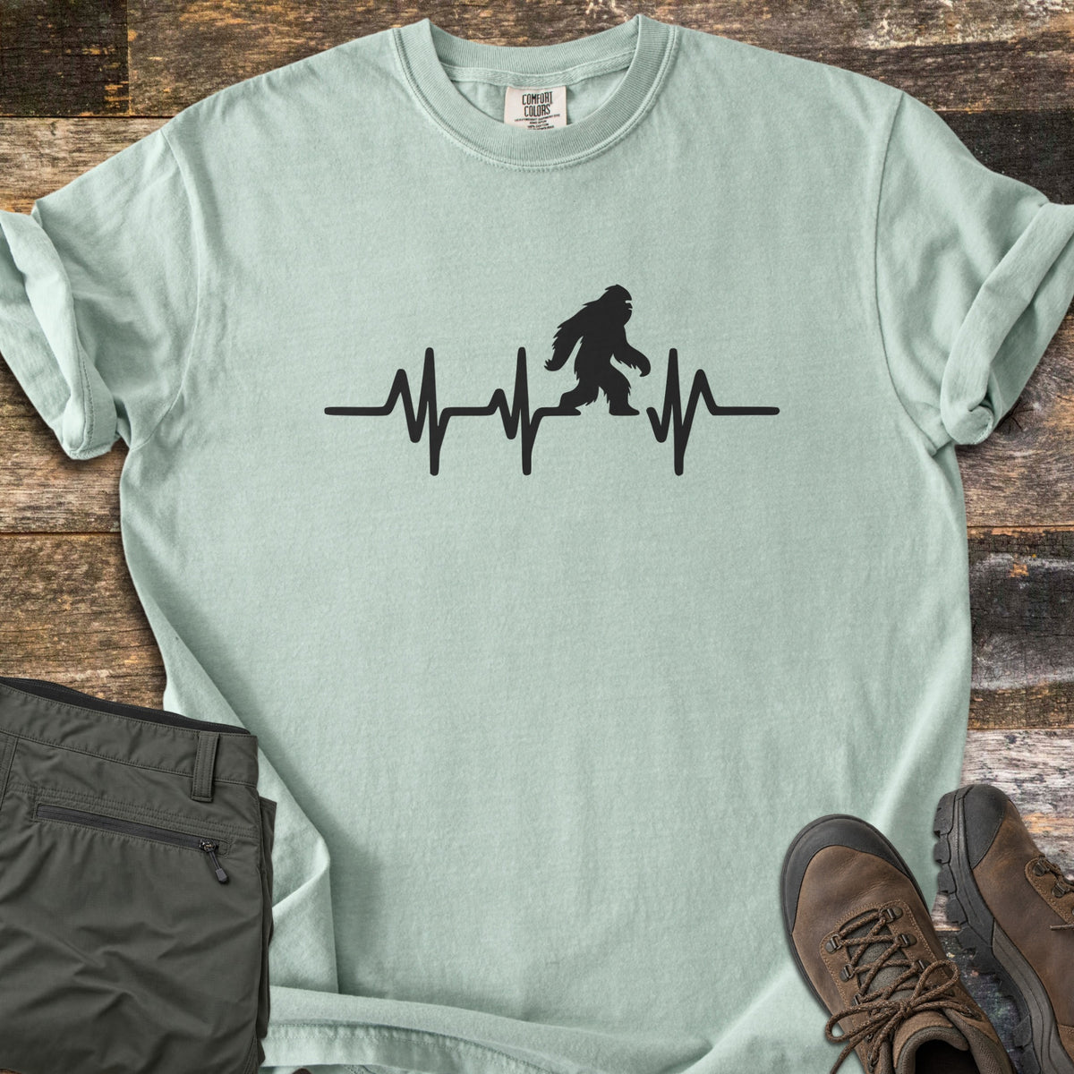 Bigfoot Heartbeat Comfort Colors T-shirt in Bay, laid flat. A black heartbeat line runs across the chest with a walking Bigfoot silhouette in the middle; there are no words. The shirt lies on weathered wood beside folded olive hiking pants and brown hiking boots.