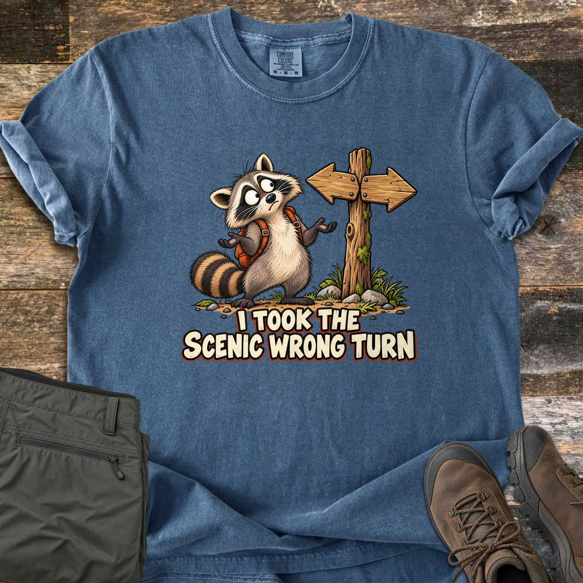 I Took the Scenic Wrong Turn Comfort Colors T-shirt in Denim, laid flat. A cartoon raccoon wearing an orange backpack shrugs beside a wooden signpost with arrows pointing both ways, above the words I Took the Scenic Wrong Turn. Olive hiking pants and brown hiking boots sit on weathered wood.