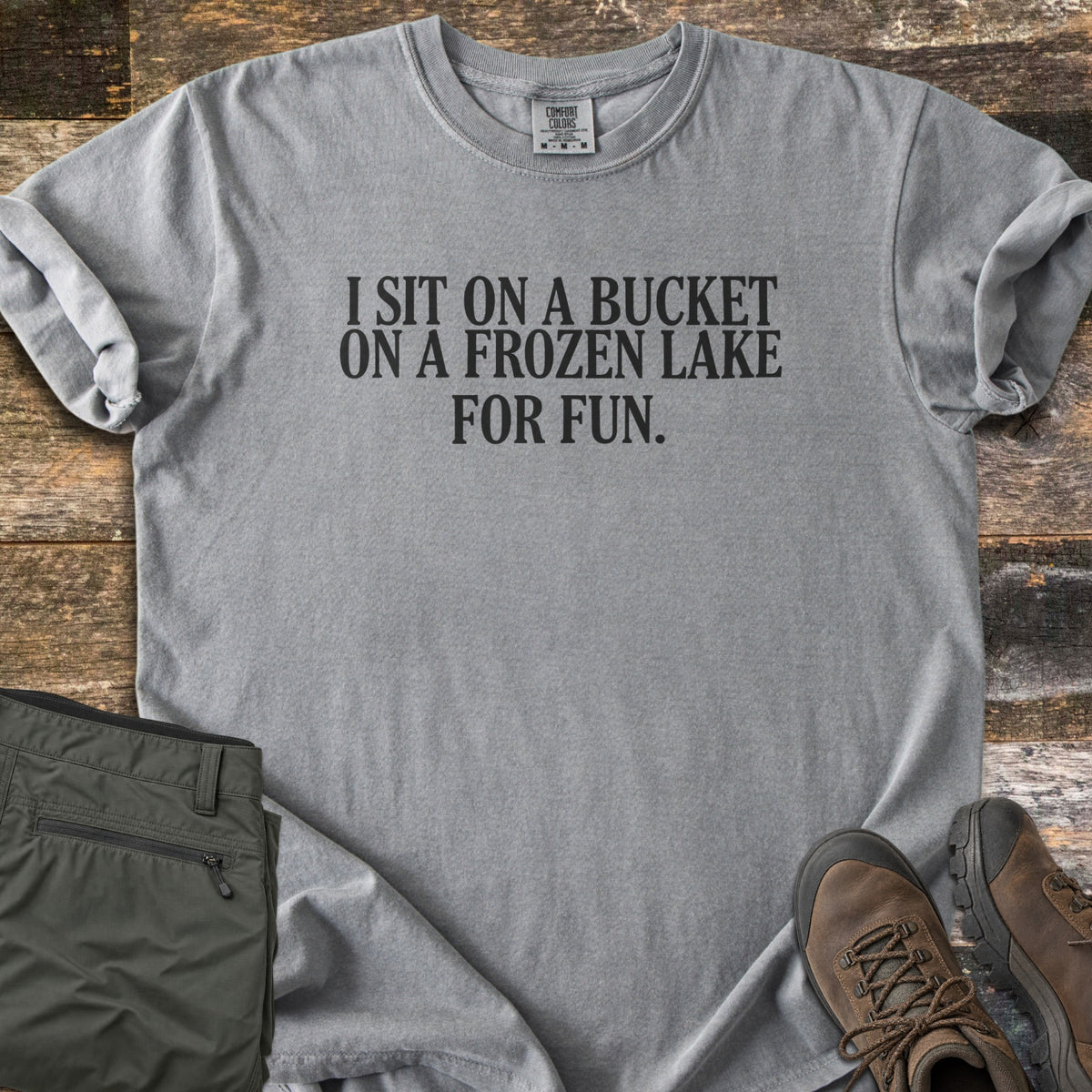Bucket On A Frozen Lake Comfort Colors T-shirt in Grey, laid flat. Black serif text reads I SIT ON A BUCKET ON A FROZEN LAKE FOR FUN. The shirt has rolled sleeves and lies on weathered wood planks beside folded olive hiking pants and brown hiking boots.