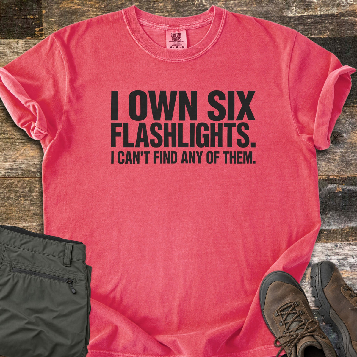 Six Flashlights Comfort Colors T-shirt in Watermelon, laid flat. Bold black text reads I OWN SIX FLASHLIGHTS. I CAN'T FIND ANY OF THEM. The shirt has rolled sleeves and lies on weathered wood beside folded olive cargo pants and brown hiking boots.