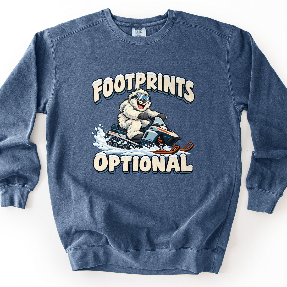 Footprints Optional Comfort Colors crewneck sweatshirt in Denim, laid flat. A cartoon yeti in ski goggles grins while riding a snowmobile through spraying snow, between the words Footprints Optional in cream letters topped with snow. Shown on a white background.