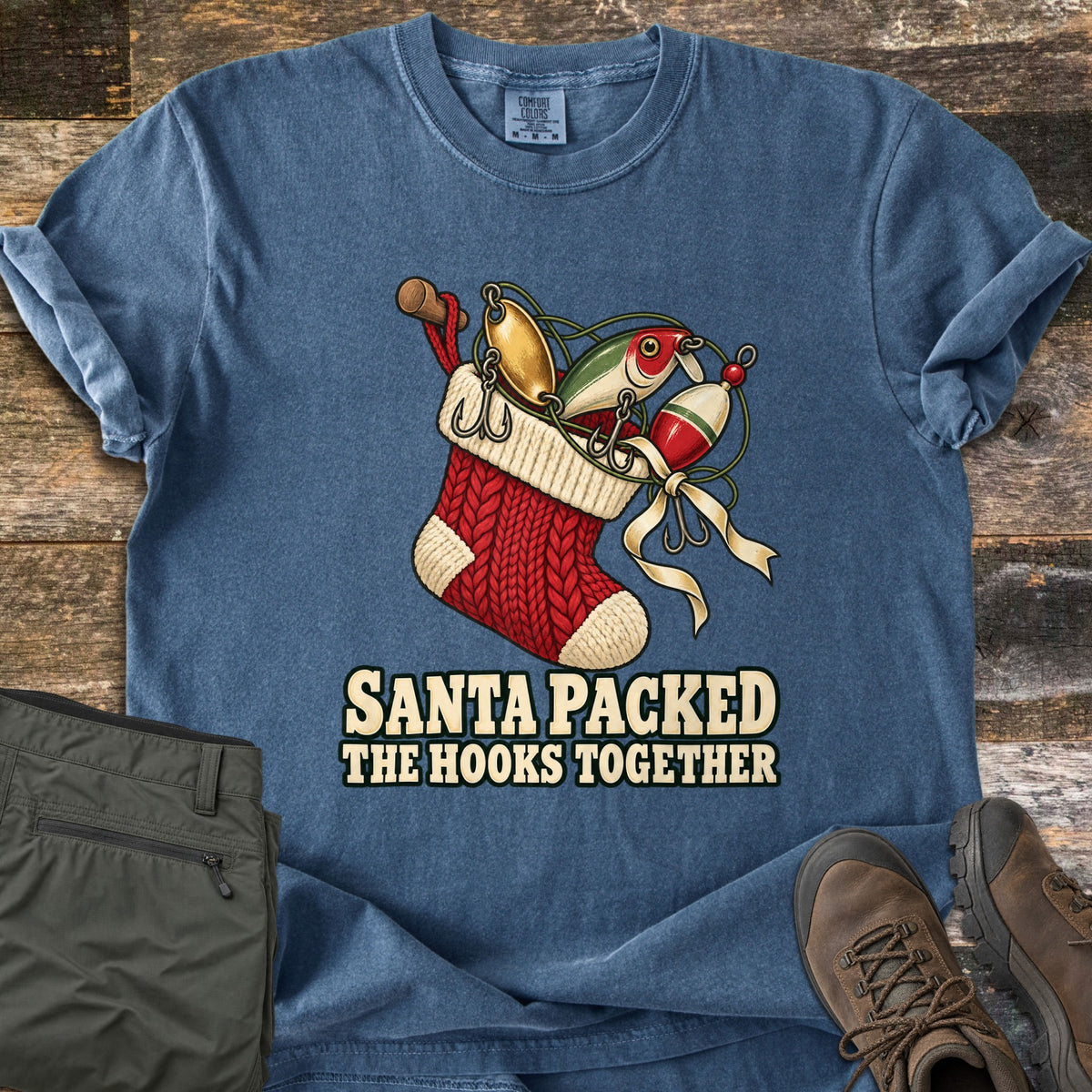 Santa Packed The Hooks Together Comfort Colors T-shirt in Denim, laid flat. A red knit Christmas stocking holds fishing lures, a bobber, and a reel with a cream ribbon, above the words Santa Packed The Hooks Together. Olive pants and brown boots sit beside it on weathered wood.