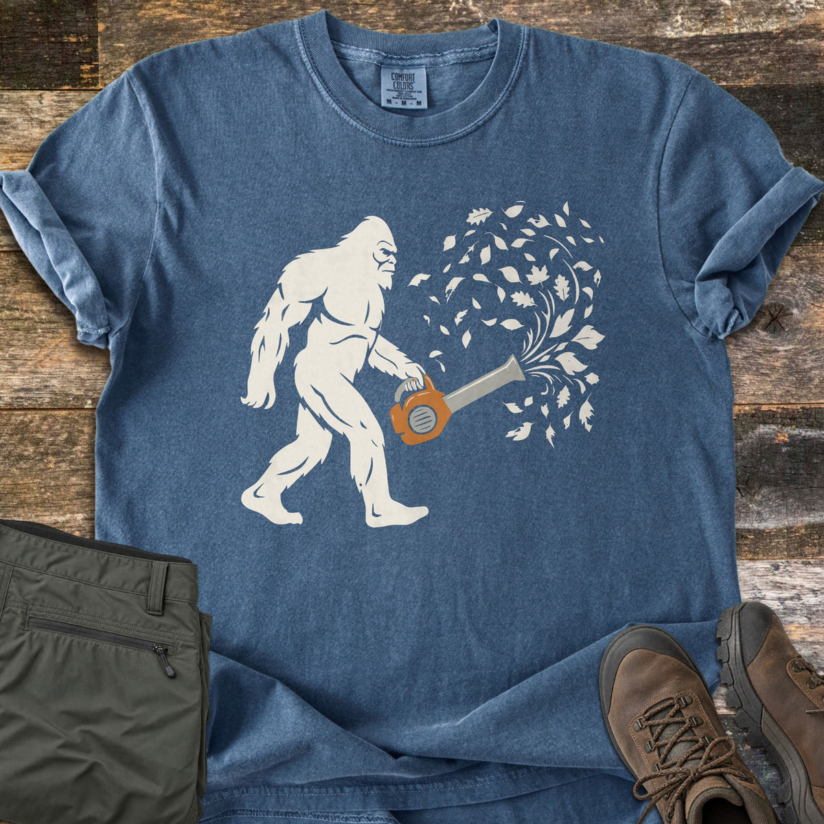 Bigfoot Leaf Blower Comfort Colors T-shirt in Denim, laid flat. A cream silhouette of Bigfoot walks while holding an orange leaf blower that sends a swirl of cream leaves into the air. Olive pants and brown hiking boots sit beside it on weathered wood planks.