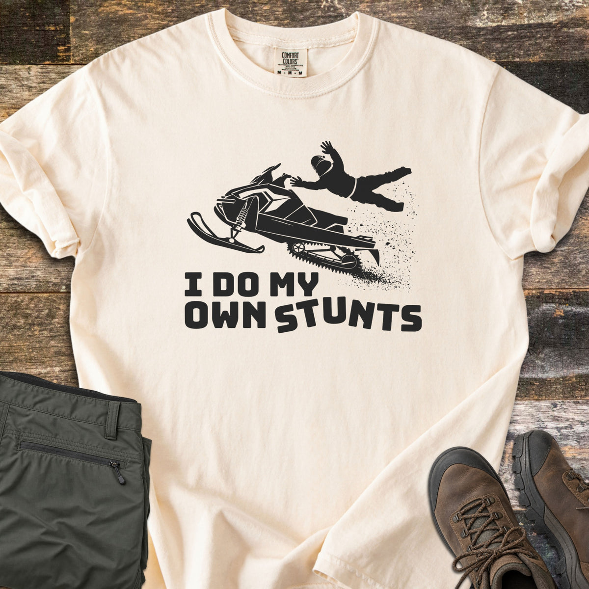 I Do My Own Stunts Comfort Colors T-shirt in Ivory, laid flat. A black silhouette of a rider flying off a jumping snowmobile, snow spraying behind, above the words I Do My Own Stunts. Folded olive cargo pants and brown hiking boots sit beside it on weathered wood.