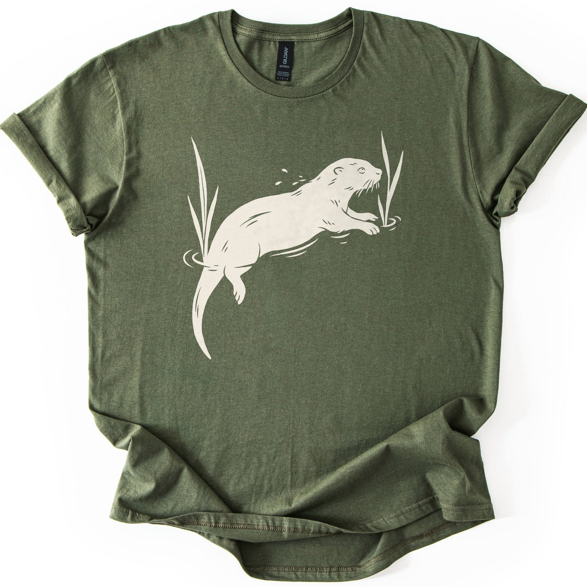 Swimming Otter Comfort Colors T-shirt in Military Green, laid flat. A cream linocut-style otter swims through rippling water between tall reeds, with droplets above its head. No words are printed. The sleeves are cuffed and the shirt lies on a white background.