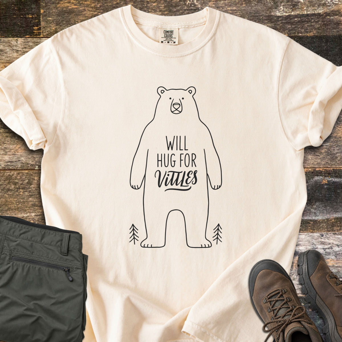 Will Hug For Vittles Comfort Colors T-shirt in Ivory, laid flat. A black outline of a standing bear has the words Will Hug For Vittles on its belly, with two small pine trees by its feet. Folded olive hiking pants and brown hiking boots sit beside it on weathered wood.