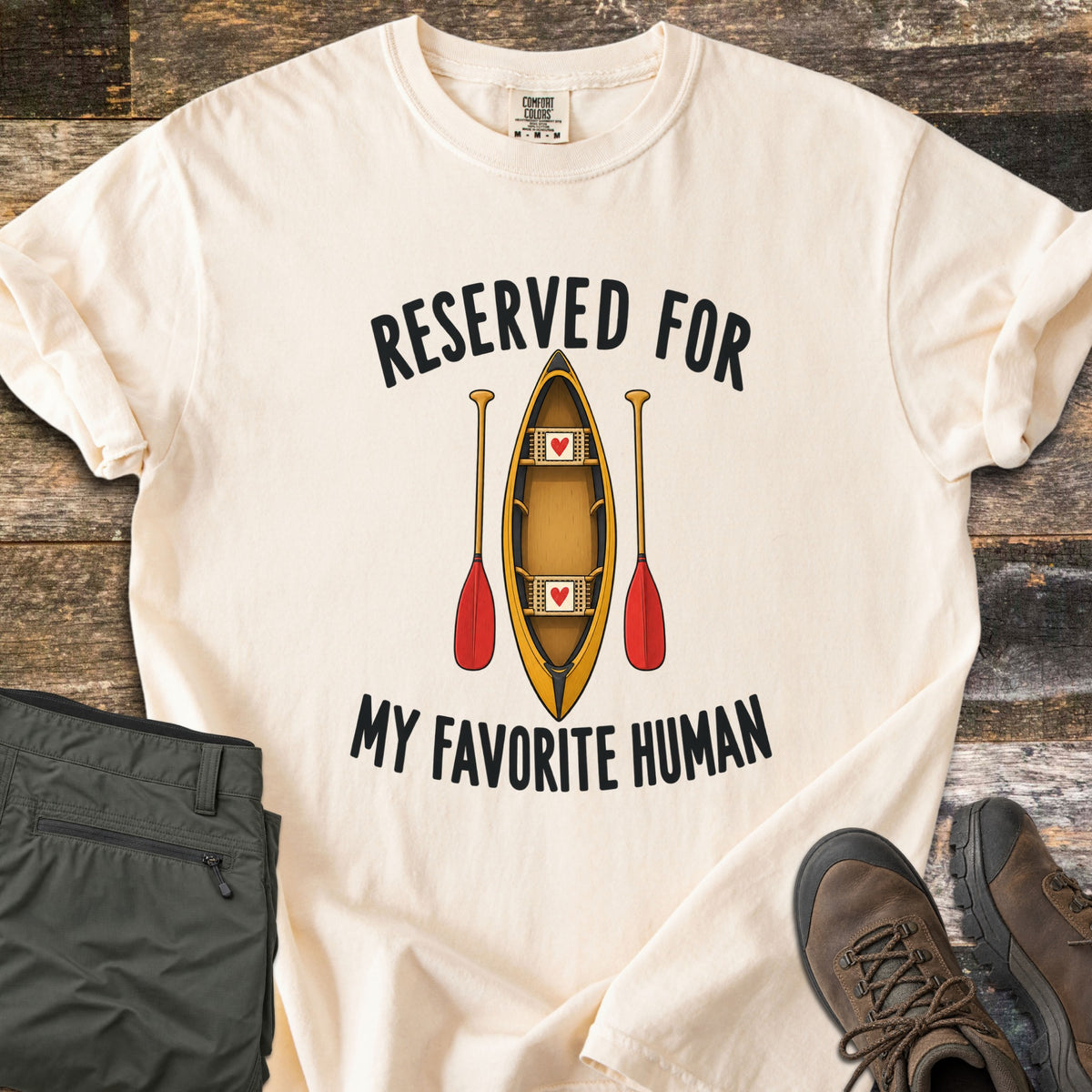 Reserved For My Favorite Human Comfort Colors T-shirt in Ivory, laid flat. A yellow canoe seen from above with a red heart on each seat sits between two red-bladed paddles, under the words Reserved For and above My Favorite Human. Olive pants and brown hiking boots lie beside it on wood planks.