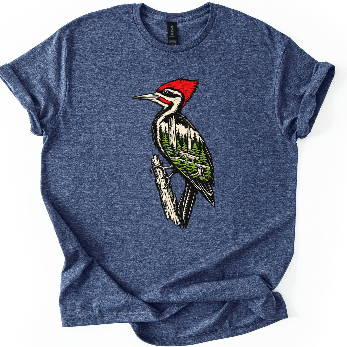 Woodpecker Forest Comfort Colors T-shirt in Heather Navy, laid flat. A red-crested woodpecker clings to a weathered branch, its black and cream body filled with pine trees and fallen logs. No words are printed. The sleeves are cuffed and the shirt lies on a white background.