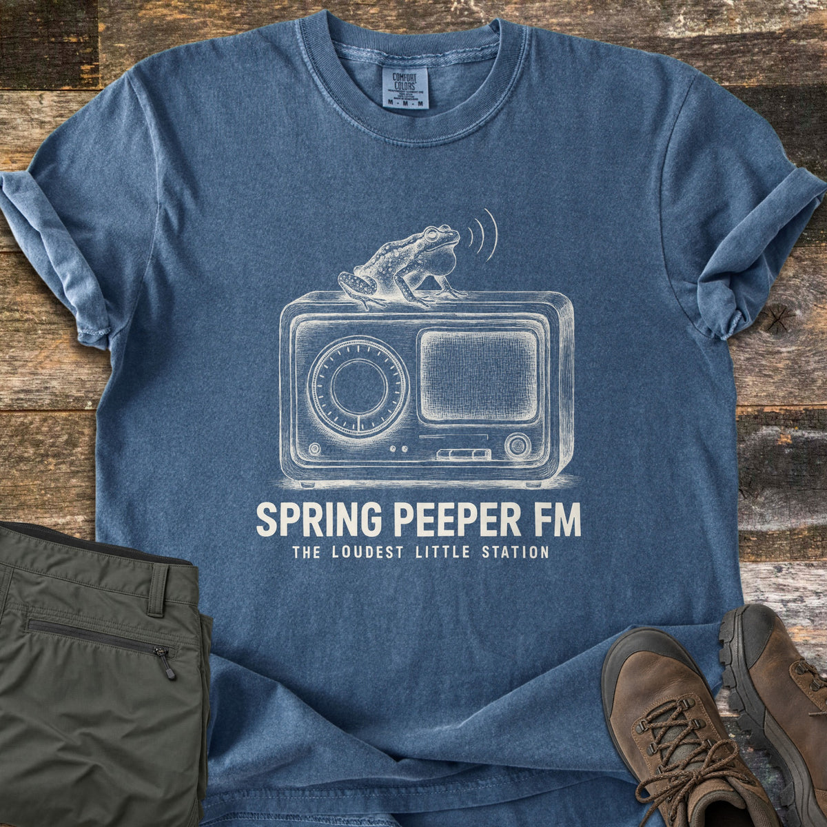 Spring Peeper FM Comfort Colors T-shirt in Denim, laid flat. A white sketch of a frog with puffed throat sitting atop a vintage radio, sound waves above, over the words Spring Peeper FM, The Loudest Little Station. On weathered wood with green pants and brown hiking boots.