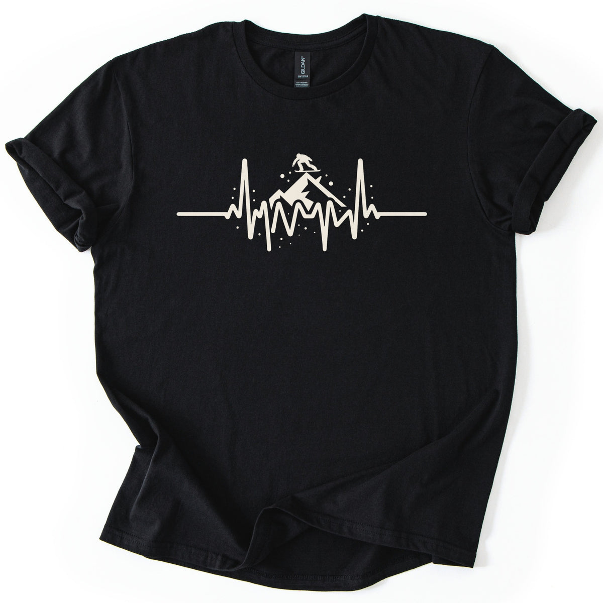 Snow Boarder Heartbeat Comfort Colors T-shirt in Black, laid flat. A cream heartbeat line rises into a snowy mountain peak with a snowboarder silhouette riding on top, surrounded by snow dots. No words. The shirt has rolled sleeves and lies on a white background.