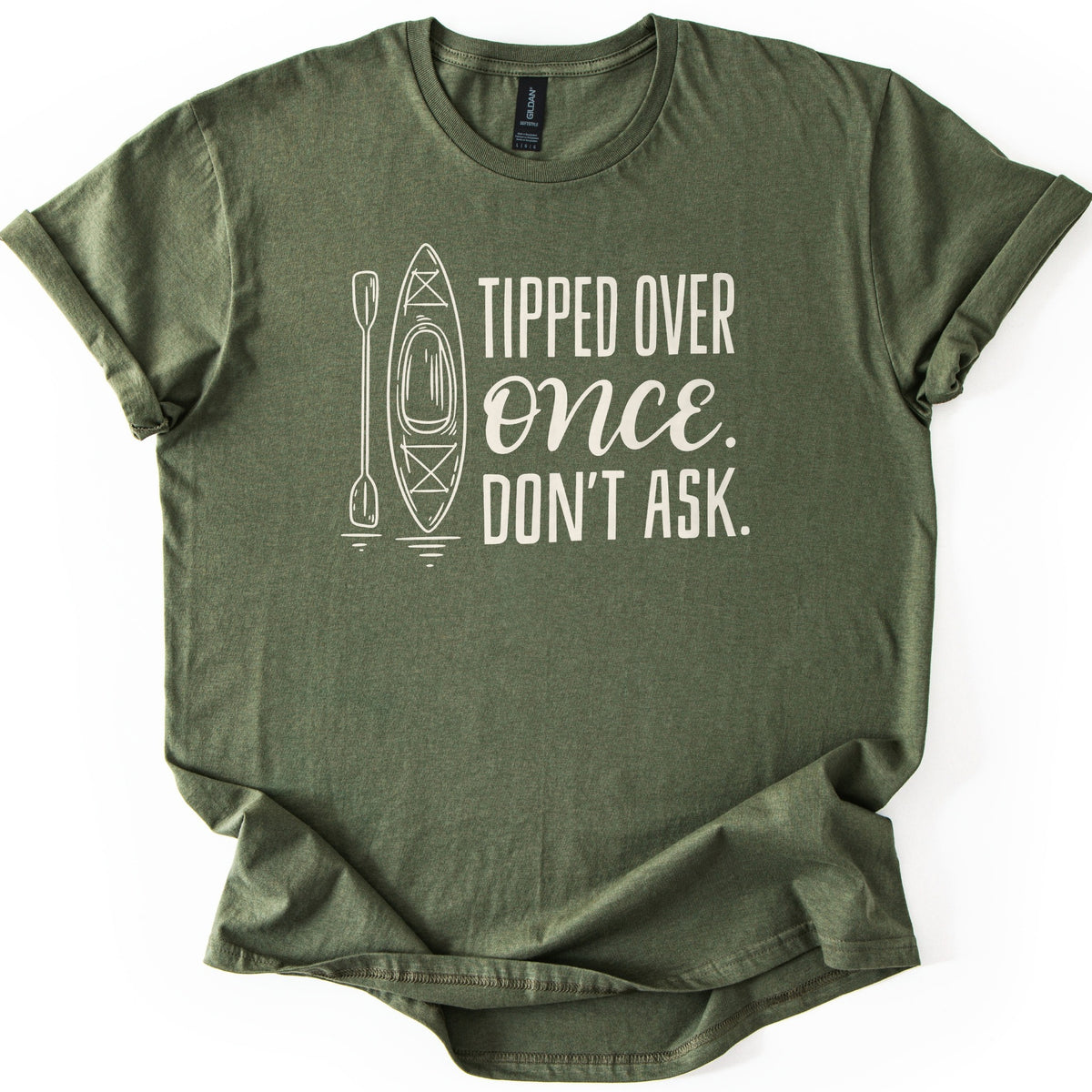 Tipped Over Once Kayak Comfort Colors T-shirt in Military Green, laid flat. A cream line drawing of an upright kayak and paddle with water ripples sits beside the words Tipped Over Once. Don't Ask., with Once in script. The shirt has cuffed sleeves on a white background.
