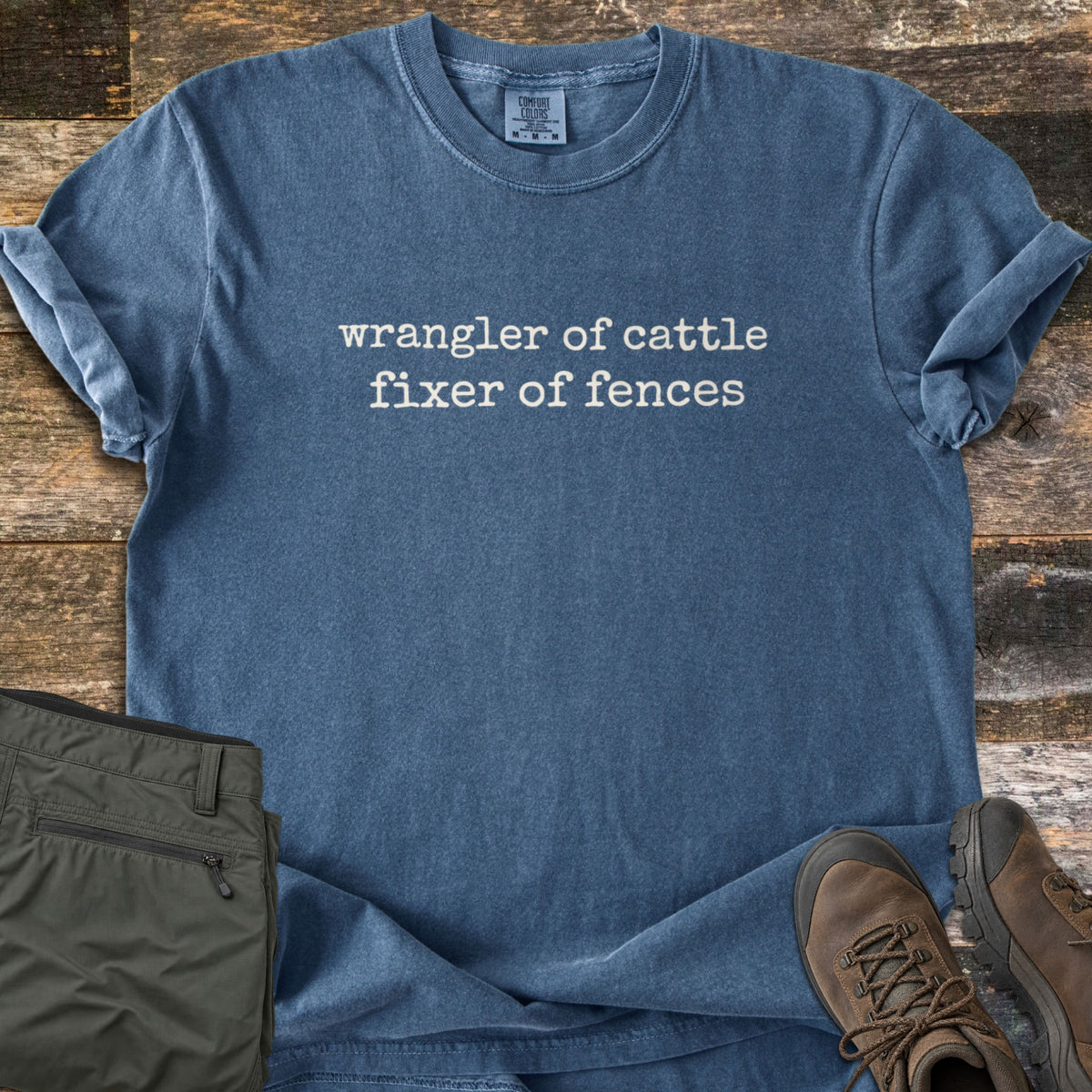 Fixer of Fences Comfort Colors T-shirt in Denim, laid flat. Small white typewriter-style text across the chest reads wrangler of cattle fixer of fences. Sleeves are cuffed; olive cargo pants and brown leather hiking boots sit beside it on weathered wood planks.