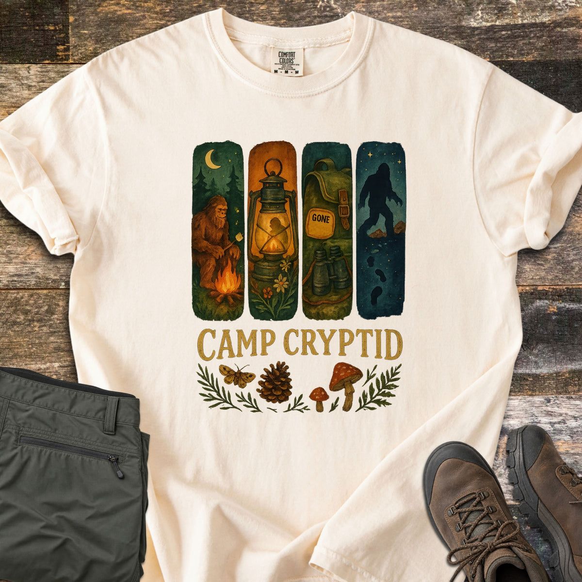 Camp Cryptid Big Foot Comfort Colors T-shirt in Ivory, laid flat. Four painted panels show Bigfoot at a campfire, a lantern, a backpack marked GONE with binoculars, and Bigfoot under stars, above CAMP CRYPTID, a moth, pinecone and mushrooms. Olive pants and hiking boots lie on wood.