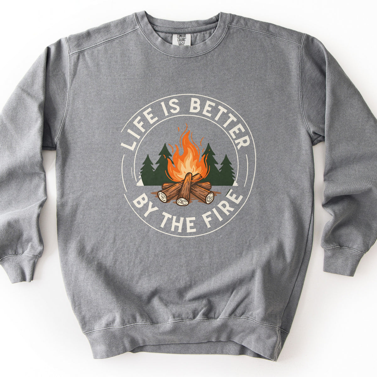 Life Is Better By The Fire Ring Comfort Colors crewneck sweatshirt in Grey, laid flat. A campfire of stacked logs with orange flames sits in front of dark green pines inside a cream ring, with the words Life Is Better By The Fire curved around it. It lies on a white surface.