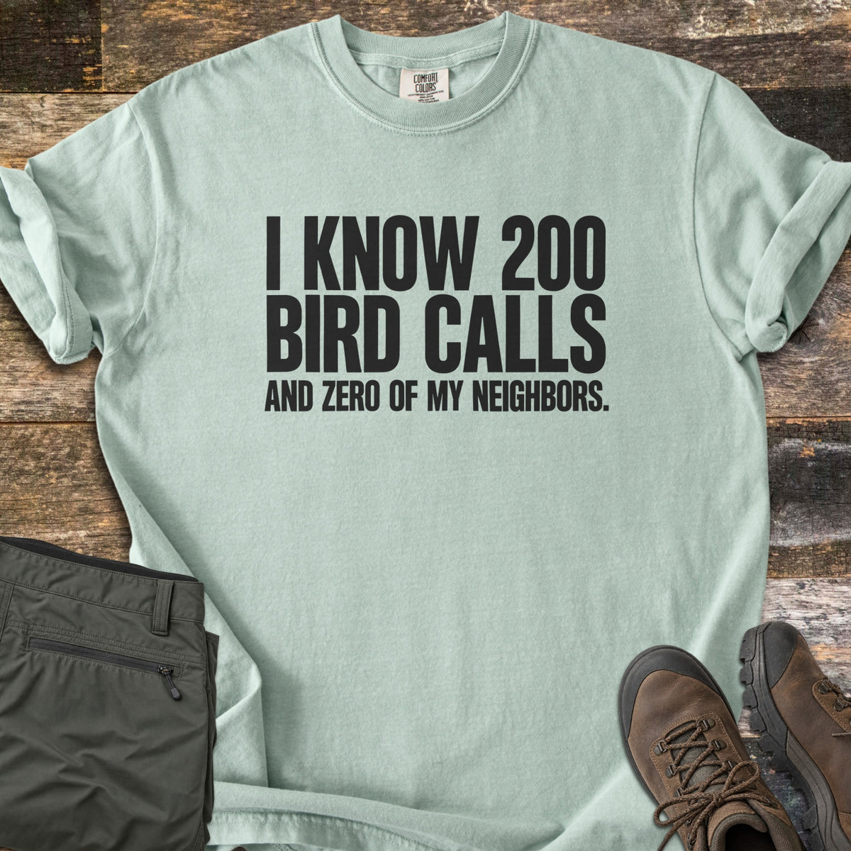 I Know 200 Bird Calls and Zero of My Neighbors Comfort Colors T-shirt in Bay, laid flat. Bold black capital letters read I KNOW 200 BIRD CALLS AND ZERO OF MY NEIGHBORS. The shirt has rolled sleeves and lies on weathered wood boards beside folded olive hiking pants and brown hiking boots.