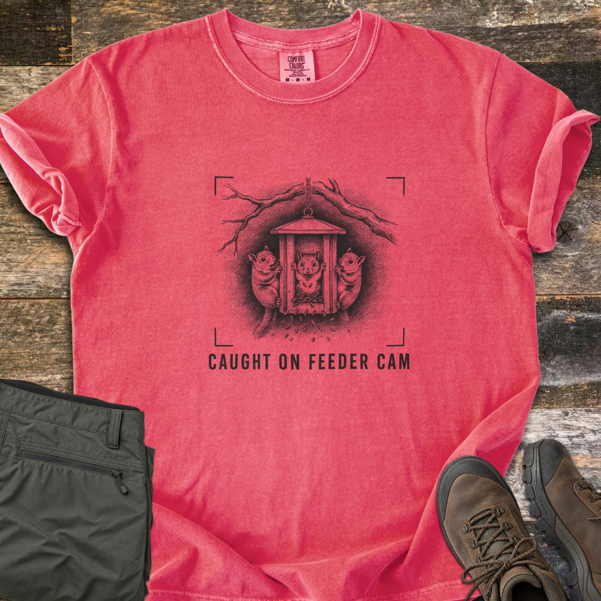 Caught On Feeder Cam Comfort Colors T-shirt in Watermelon, laid flat. A black ink sketch of three squirrels raiding a hanging wooden feeder inside camera viewfinder corners, above the words CAUGHT ON FEEDER CAM. Folded olive pants and brown hiking boots sit on weathered wood planks.